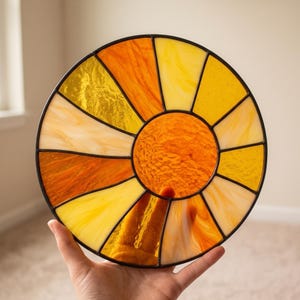 May include: A circular stained glass sun catcher featuring a central orange textured glass circle surrounded by radiating segments of yellow, amber, and orange glass. The individual glass pieces are joined together by thin black metal solder lines.