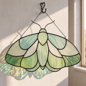 May include: A stained glass moth suncatcher featuring various shades of green and white glass panels held together by dark metal cames. The moth is suspended by two thin metal chains attached to a single dark metal hook mounted on the wall.