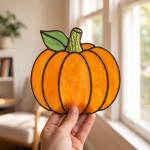 May include: A stained glass pumpkin suncatcher featuring vibrant orange marbled glass panels with dark leaded solder lines, topped with a textured light green glass leaf and a small, vertically patterned green stem, held up against a bright window.