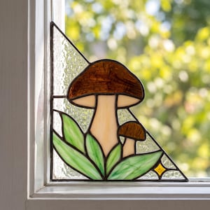 May include: A triangular stained glass window panel featuring two brown mushrooms with cream-colored stems, surrounded by green leaves and set against a textured clear glass background with a small yellow star accent in the corner.