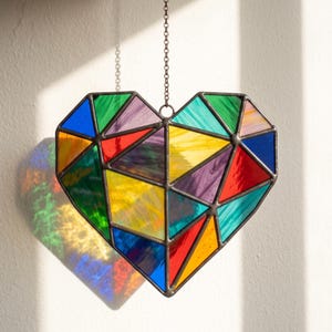 May include: A heart-shaped stained glass suncatcher composed of numerous triangular and polygonal glass panels in vibrant shades of red, blue, yellow, green, purple, and orange, held together by dark metal soldering and suspended by a metal chain.