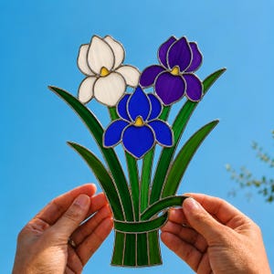 May include: A stained glass suncatcher featuring three iris flowers in white, deep blue, and purple, with green stems and leaves bound together at the base. The piece is constructed with soldered metal lines separating each colored glass segment.