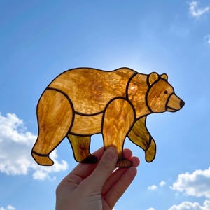 May include: A stained glass suncatcher shaped like a walking bear, crafted from amber-coloured glass with a marbled texture and dark solder lines outlining the body segments, held up against a bright blue sky with scattered white clouds.