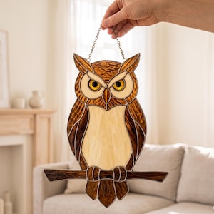 May include: A stained glass owl suncatcher featuring textured brown and cream glass panels with yellow eyes. The owl is perched on a horizontal branch and is suspended by a thin metal chain, showcasing intricate lead lines and detailed craftsmanship.