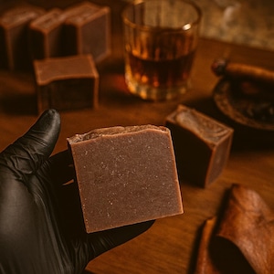 May include: A brown soap bar is held in a black-gloved hand. Several other bars are visible, along with a glass of amber liquid, a lit cigar, and a small dish. The scene suggests a masculine theme.