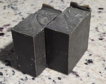 Puede incluir: Dos barras rectangulares de jabón gris oscuro con un patrón veteado en la parte superior. Las barras de jabón están colocadas una al lado de la otra sobre una superficie moteada blanca y gris.