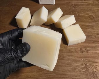 May include: A collection of handmade soap bars in various shapes and sizes. The image shows a large rectangular bar being held, along with several smaller, angled bars and a packaged bar. The soap has a creamy, off-white color and is displayed on a wooden cutting board.
