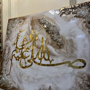 May include: A decorative wall art piece featuring gold Arabic calligraphy on a white and brown marbled background. The artwork is framed with silver and gold accents, creating a luxurious aesthetic.