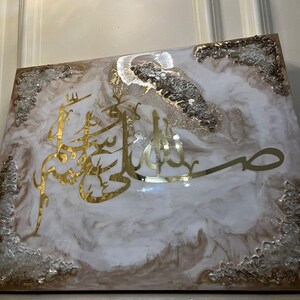 May include: A square decorative piece with a white and brown marbled design. Gold Arabic calligraphy is featured prominently in the center. The edges are adorned with clear and gold-colored embellishments.