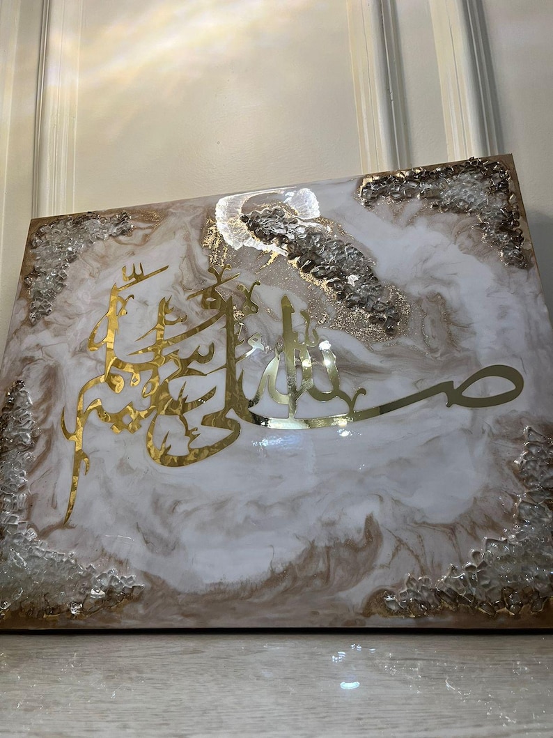 May include: A square decorative piece with a white and brown marbled design. Gold Arabic calligraphy is featured prominently in the center. The edges are adorned with clear and gold-colored embellishments.