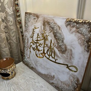 May include: A square canvas art piece with gold Arabic calligraphy on a white and gold marbled background. The edges are trimmed with gold and silver accents. The artwork is displayed on a light-colored wooden surface.