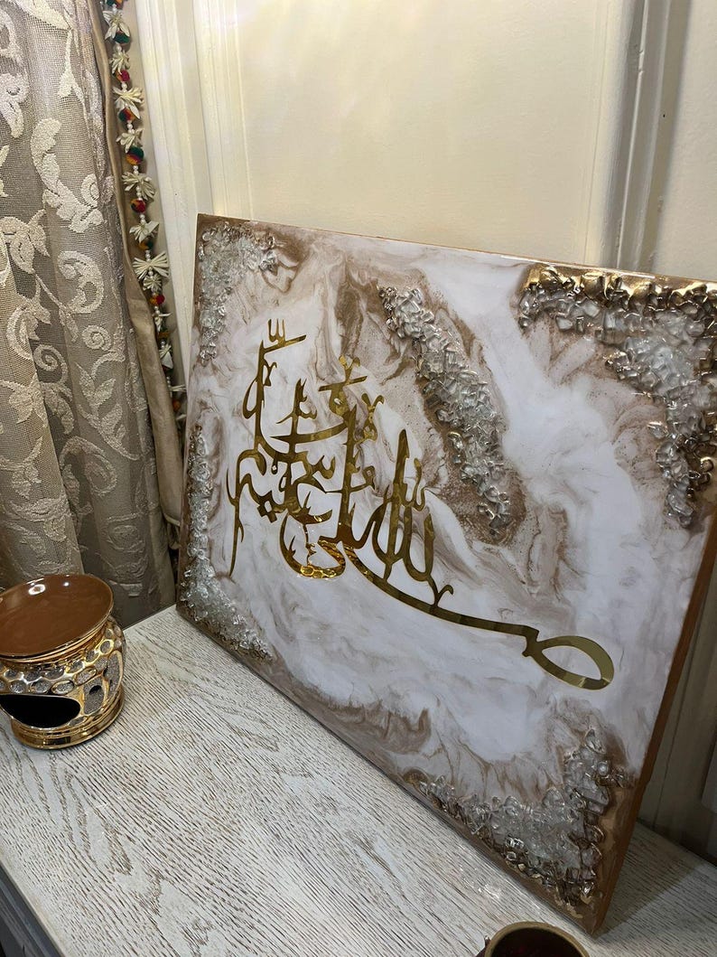 May include: A square canvas art piece with gold Arabic calligraphy on a white and gold marbled background. The edges are trimmed with gold and silver accents. The artwork is displayed on a light-colored wooden surface.