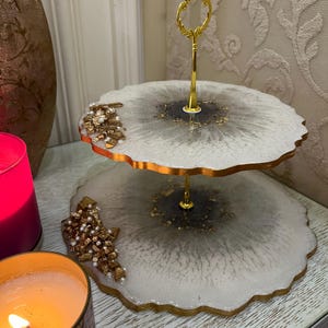May include: Two-tiered serving tray with a gold-colored handle. The tray has a white and gray marbled design with gold accents and a gold-colored trim. Decorative gold and pearl clusters adorn the edges. Candles are in the foreground.