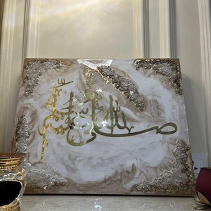 May include: A rectangular artwork featuring gold Arabic calligraphy on a white and beige marbled background. The edges are adorned with clear, textured embellishments. The piece is displayed on a light-colored surface.