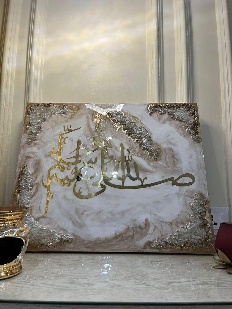 May include: A rectangular artwork featuring gold Arabic calligraphy on a white and beige marbled background. The edges are adorned with clear, textured embellishments. The piece is displayed on a light-colored surface.