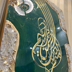 May include: A decorative artwork featuring gold Arabic calligraphy on a dark green background. The artwork is framed with gold and clear crystal-like accents. The calligraphy is in a stylized script.