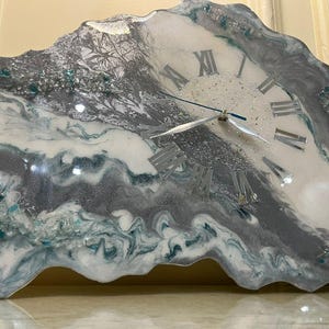 May include: A decorative wall clock with a unique, organic shape. The clock face features Roman numerals and silver hands. The clock's surface has a marbled effect with shades of gray, white, and teal, and is accented with sparkling details.