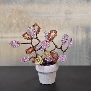 May include: A miniature beaded flower arrangement in a small, light gray pot. The flowers are crafted from translucent pink, yellow, and amber beads, with dark brown wire stems. The pot sits on a dark surface.