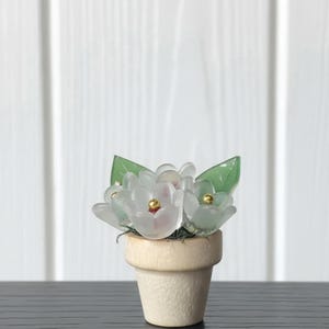 May include: A miniature potted plant featuring frosted white glass flowers with small gold-toned centers and smooth green glass leaves. The flowers are arranged in a small, light-colored wooden pot with a textured, natural finish and a wide rim.