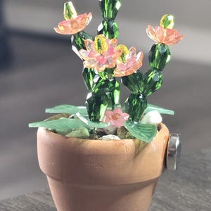 May include: A miniature cactus figurine crafted from faceted green and pink crystal beads, potted in a small terracotta-colored clay pot. The cactus features multiple stems topped with pink and yellow crystal flowers, with light green leaves.