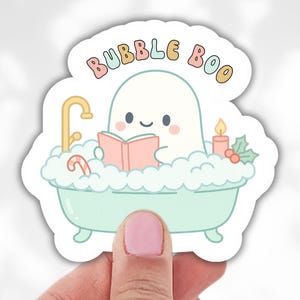 May include: A sticker featuring a cute ghost character in a bathtub, reading a book. The sticker has the words "Bubble Boo" at the top. The ghost is in a mint green bathtub with bubbles, a candy cane, and a candle. The sticker is held by a hand.