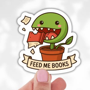Puede incluir: Una pegatina con una planta de dibujos animados sonriente, comiendo un libro rojo. La planta está en una maceta marrón, con una pancarta que dice "FEED ME BOOKS". La pegatina está sujeta por una mano.