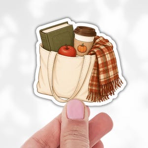 May include: A cream-colored canvas tote bag containing a green hardcover book, a red apple, and a disposable coffee cup with a pumpkin illustration. A red, orange, and white plaid scarf with fringe hangs over the side of the tote bag.