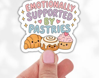 May include: A die-cut sticker featuring the text EMOTIONALLY SUPPORTED BY PASTRIES in pink, purple, green, and blue. Below the text are three cartoon pastries with faces: a golden brown croissant, a cinnamon roll, and a pink frosted cupcake.