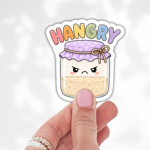 May include: A die-cut sticker featuring a cartoon glass jar with a beige textured filling, a purple floral lid, and a grumpy facial expression. Above the jar, the word HANGRY is written in colorful, pastel-toned block letters. The sticker has a white border.