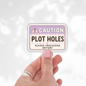 May include: A rectangular sticker with a light purple and cream background featuring a pink triangle with an exclamation point. The text reads CAUTION PLOT HOLES and READER PROCEEDING ANYWAY in dark brown and black lettering with a small pink star.
