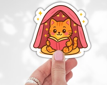 May include: A sticker featuring a cartoon orange tabby cat reading a book under a pink tent. The cat is sitting on a yellow cushion, and the tent is decorated with small yellow stars. The sticker has a white border.