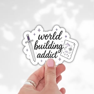 May include: A white sticker with the words "world building addict" in black script. The sticker features a sword, a map, and purple star accents. The sticker is held by a hand with pink nail polish and silver rings.