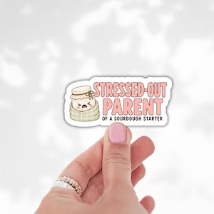 May include: A die-cut sticker featuring a cartoon jar with a distressed face wrapped in a beige blanket, accompanied by the text STRESSED-OUT PARENT OF A SOURDOUGH STARTER in pink and black lettering against a white background.