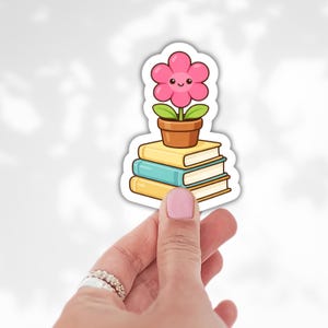 May include: A sticker featuring a smiling pink flower in a brown pot atop a stack of three books. The books are yellow and teal. The sticker is held by a hand with pink nail polish.