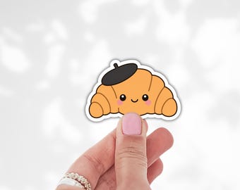 May include: A die-cut sticker featuring a golden-brown croissant with a smiling face, rosy cheeks, and a small black beret perched on top. The illustration has a thick white border and a clean, simple design with bold outlines and flat colors.