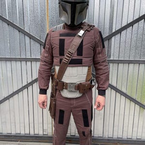 Puede incluir: Un disfraz de Mandaloriano marr&oacute;n y negro, que incluye un casco gris met&aacute;lico, una armadura para el pecho, pantalones y un cintur&oacute;n utilitario con bolsillos. El atuendo tiene detalles en negro. El casco es el punto central.