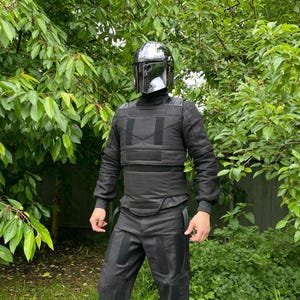 Puede incluir: Un conjunto de armadura negra mandaloriana, que incluye un casco con visera reflectante, una parte superior de manga larga y pantalones. La armadura tiene un dise&ntilde;o t&aacute;ctico con paneles y correas reforzadas. La persona est&aacute; de pie frente a un fondo de hojas verdes.
