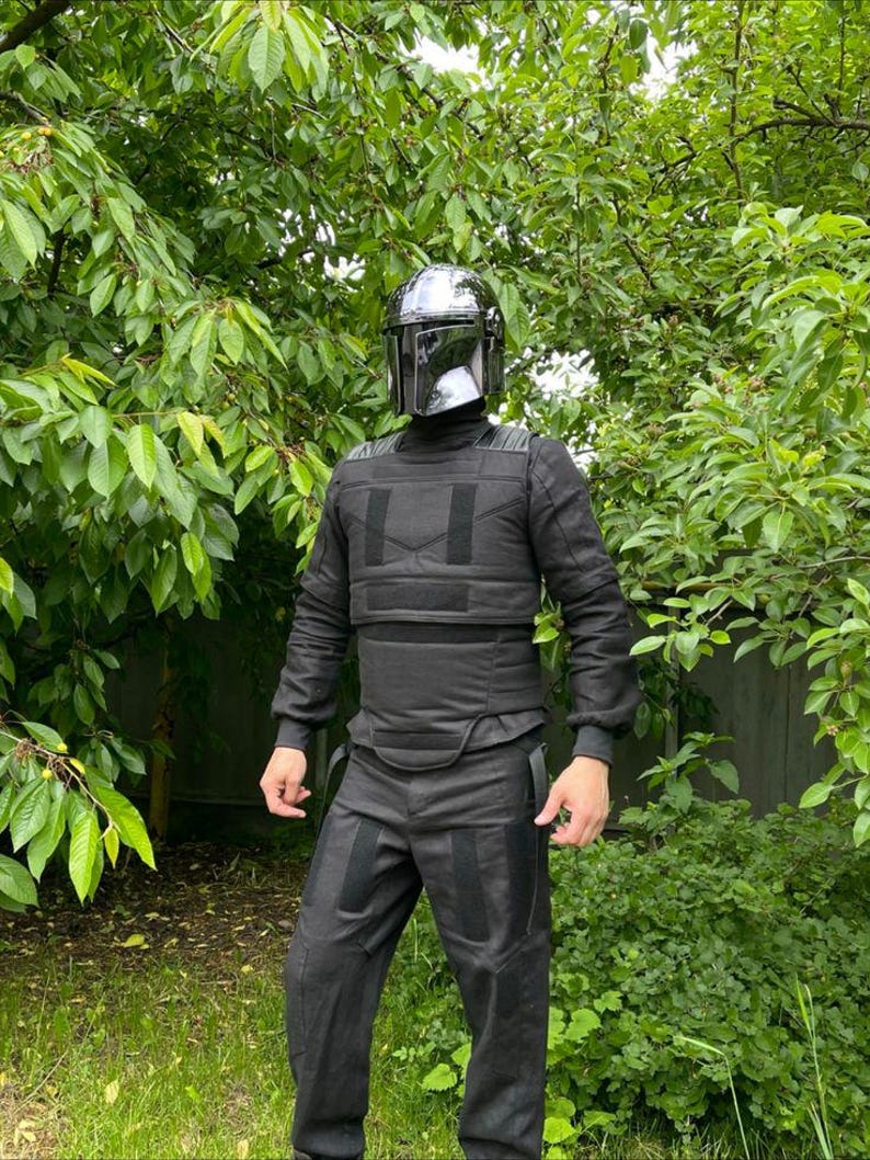 Puede incluir: Un conjunto de armadura negra mandaloriana, que incluye un casco con visera reflectante, una parte superior de manga larga y pantalones. La armadura tiene un dise&ntilde;o t&aacute;ctico con paneles y correas reforzadas. La persona est&aacute; de pie frente a un fondo de hojas verdes.