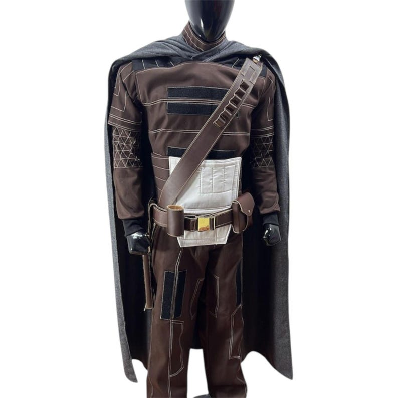 Puede incluir: Un disfraz marr&oacute;n y negro con una capa gris. El disfraz presenta una t&uacute;nica y pantalones marrones con detalles en negro, una pieza blanca en el pecho y un cintur&oacute;n de cuero marr&oacute;n con hebilla dorada. La capa es gris y cubre los hombros.