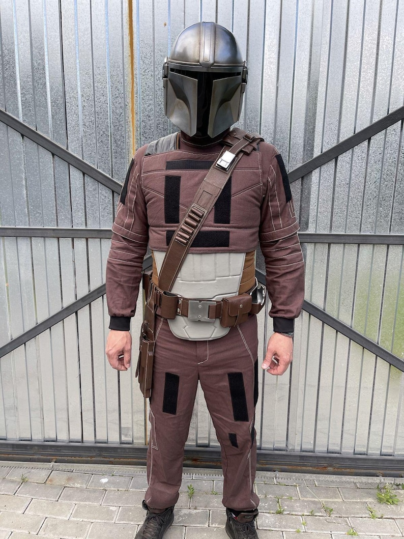 Puede incluir: Un disfraz de Mandaloriano marr&oacute;n y negro, que incluye un casco gris met&aacute;lico, una armadura para el pecho, pantalones y un cintur&oacute;n utilitario con bolsillos. El atuendo tiene detalles en negro. El casco es el punto central.