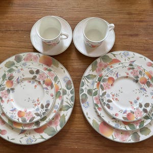 May include: A set of fine china with a floral and fruit design. The set includes two cups and saucers, two dinner plates, and two smaller plates. The plates and cups have a white background with pink flowers, peaches, and green leaves.