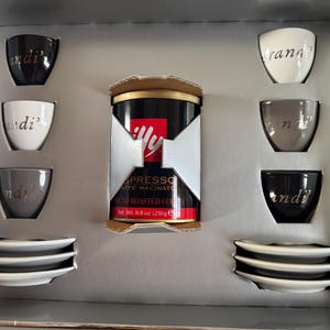 May include: An espresso coffee set with six cups in black, white, and grey, each with a silver inscription. The set includes matching saucers and a can of ground roasted coffee. The coffee can is 250g.