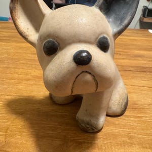 Può includere: Statuina in ceramica di un bulldog francese. Il cane ha un corpo beige con dettagli neri sulle orecchie, sugli occhi e sul naso. La statuina è seduta sulle zampe posteriori, con le zampe anteriori estese.