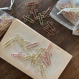 May include: A collection of cross-shaped paper clips in gold, rose gold, and copper colors. Some clips are scattered on a beige surface, while others are in small, sheer drawstring bags. The background is a dark wood grain.