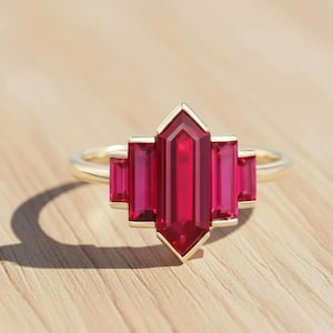 May include: A gold ring featuring a central, elongated, ruby-red gemstone flanked by four smaller, rectangular gemstones of the same color. The ring's design is geometric and modern, set against a light wooden surface.