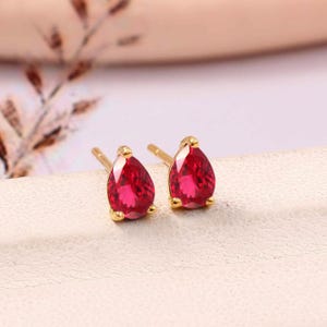 Peut inclure: Une paire de boucles d'oreilles à clous dorées, chacune ornée d'une pierre précieuse rouge vif en forme de goutte. Les boucles d'oreilles sont posées sur une surface blanche, avec un arrière-plan flou rose et doré.