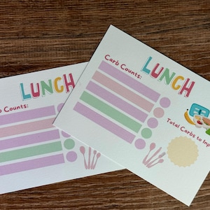 Puede incluir: Dos tarjetas blancas con la palabra "LUNCH" en letras coloridas. Las tarjetas tienen un diseño para el seguimiento de los carbohidratos, con barras y círculos de colores. También hay una pequeña ilustración de una fiambrera, un plátano y un sándwich.