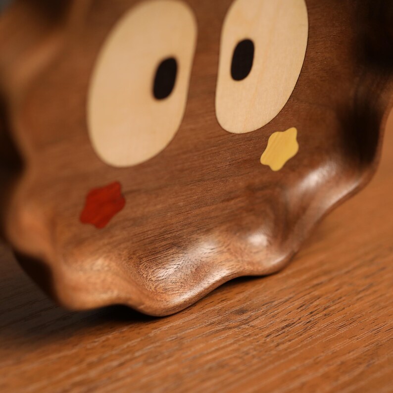 May include: A wooden decorative bowl with a cartoon face design. The bowl is brown with light tan eyes, black pupils, a red mouth, and a yellow cheek. The bowl has a wavy edge.