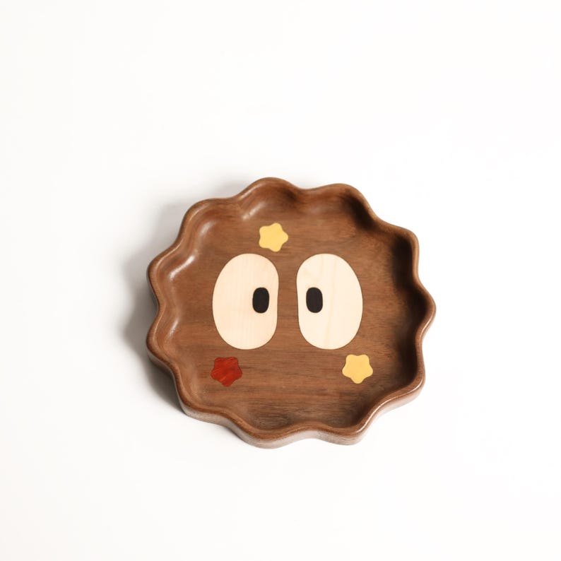 May include: A wooden tray with a whimsical design. The tray is brown with a wavy edge and features a cartoon face with large, light-colored eyes and small star accents in red and yellow. The tray is likely for decorative or functional use.