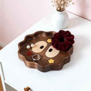 May include: A wooden tray with a whimsical design, featuring a dark brown base and inlaid light wood and colorful accents. The tray holds a burgundy scrunchie, a silver ring, and a pair of pearl earrings. A small white vase with dried flowers sits in the background.